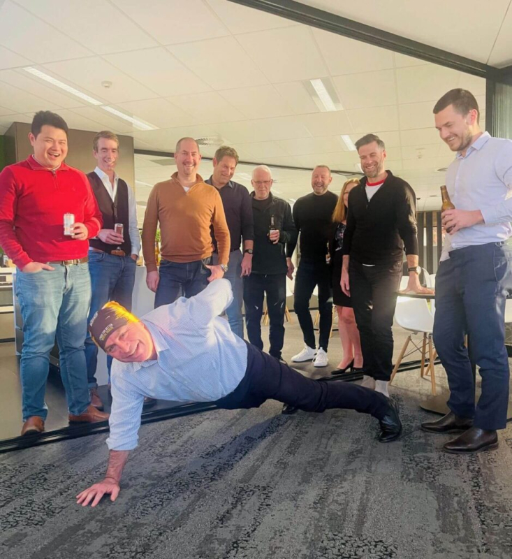 Photo of a man doing a one handed push-up with 8 people standing around him smiling.
