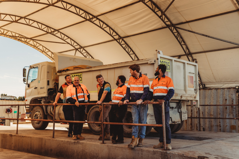 Group of 6 workman standing in a row in front of a mining truck. They are wearing hi-vis and are smiling at each other