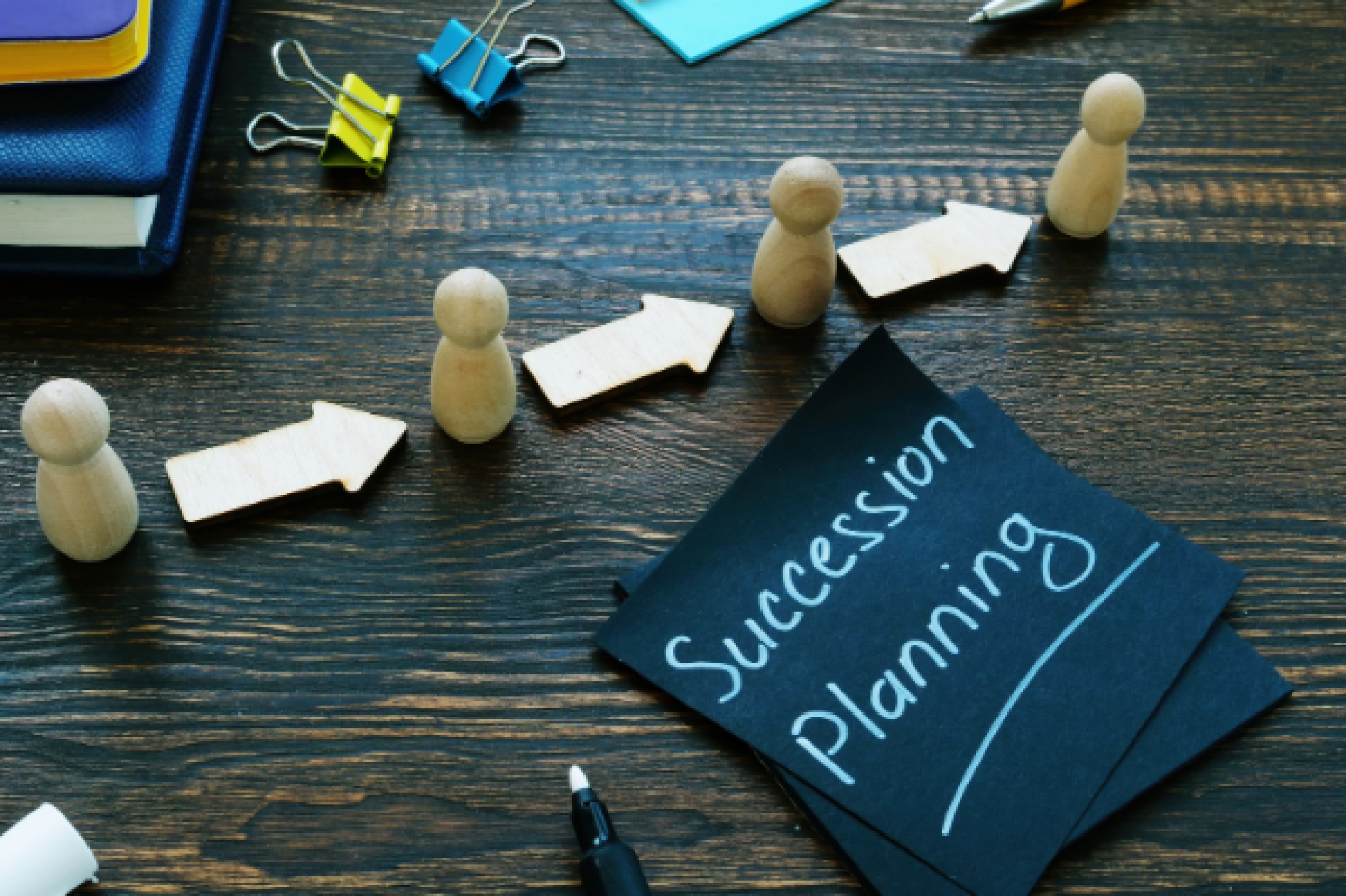 Desk with post it note with 'succession planning' written on it and arrows pointing through figurines