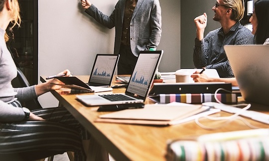 Understanding How Economic Sentiment Impacts Your Bottom Line 2 People sitting around a table in an office with a man standing pointing at a white board discussing economic sentiment